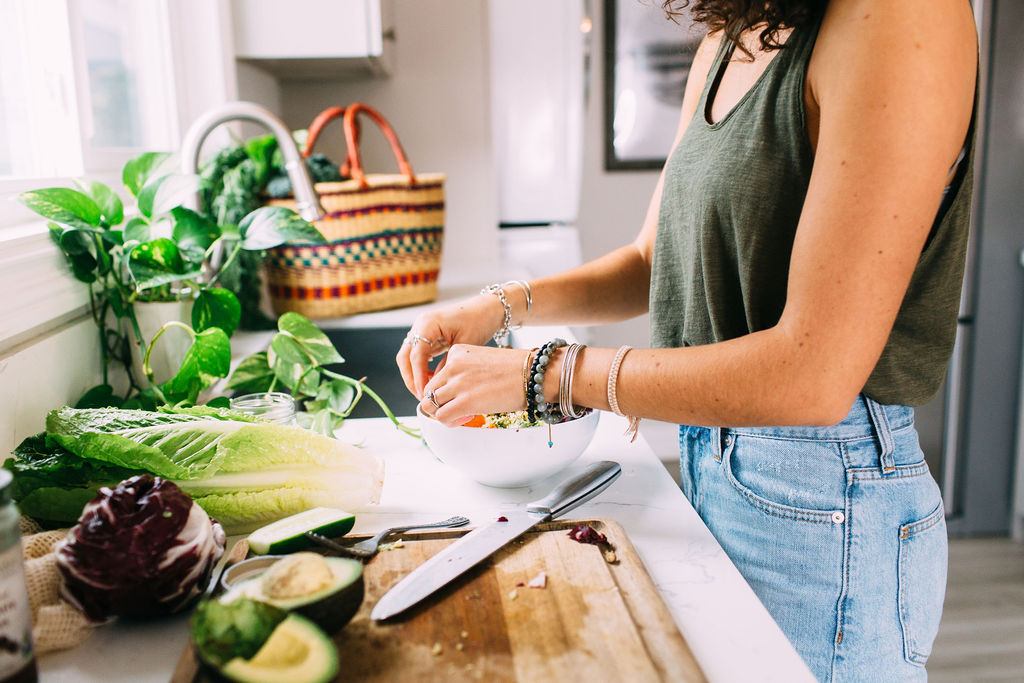 light and bright photo. View of a kitchen countertop with fresh veggies, cutting board and knife, and the sink in the background. Woman standing at the counter in jeans and an olive green tank top, prepping a  salad in a white bowl