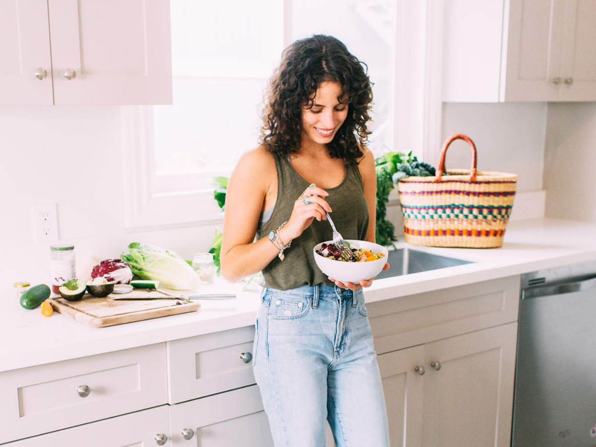 Light and airy photo. Woman with brown hair standing in the kitchen in jeans and an olive green tank top holding a white bowl of salad, smiling down at the bowl with a fork in her hand.