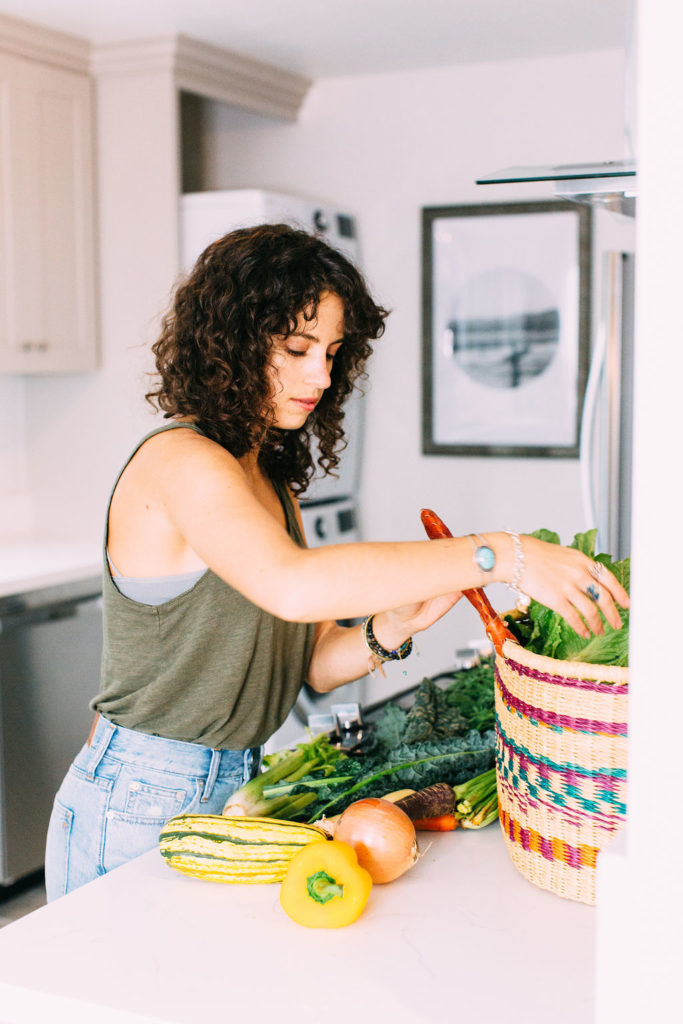 The image shows a woman in an olive green top in the kitchen, emptying a basket of produce onto the counter. There is a yellow onion, carrots, celery, kale, and delicata squash on the counter, and lettuce still in the basket.
