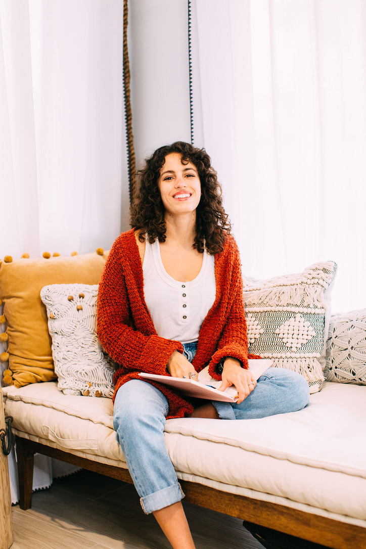 This image shows a woman sitting on a day bed, wearing light blue jeans, a white shirt and a burnt orange sweater. She has a journal in her lap and is smiling at the camera.