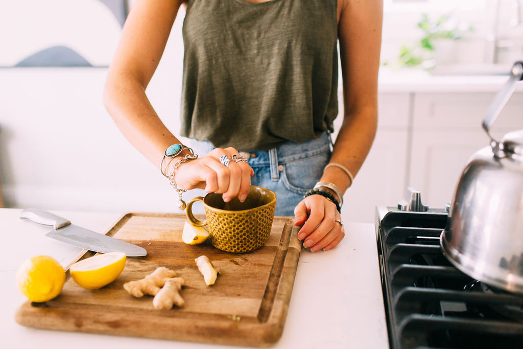 Decorative photo of a woman in an olive green tank top and light jeans at a kitchen counter, squeezing lemon into a mug. The kitchen is white and bright, and there is cut up lemon and ginger on the cutting board.