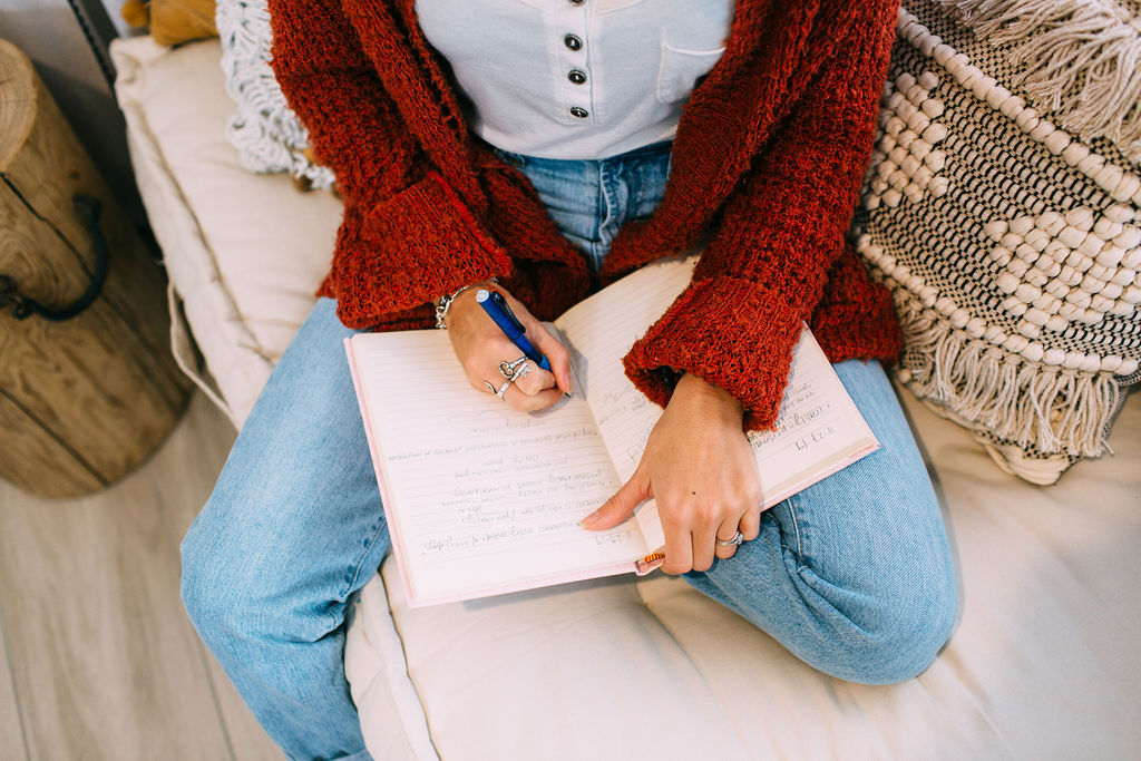 Decorative photo taken from an aerial view looking down: woman in jeans, a white shirt and a red/orange sweater sitting on a tan couch. Writing in a journal on her lap.