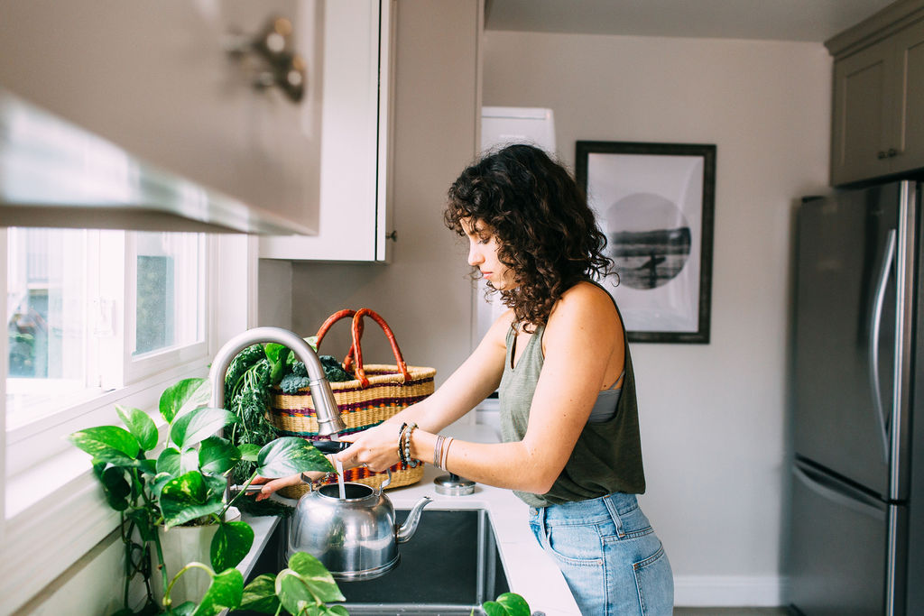 Decorative photo of a woman in a white kitchen standing at the sink, filling up a steel tea kettle with water. There is a plant in the foreground, and a basket of veggies in the background. 
