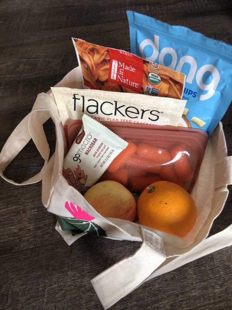 photo of foods in a tote bag: an apple, an orange, carrots in a Stasher Bag, a Go Macro bar, crackers, Dang coconut chips, and dehydrated mango